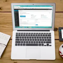 Laptop z panelem WordPress, notatnik, smartfon i filiżanka kawy na drewnianym stole.