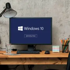 Biurko z komputerem wyświetlającym logo Windows 10 i przycisk "Zainstaluj teraz". Obok słuchawki, tablet, szkło z wodą, myszka, kredki i notatki.