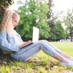 Dziewczyna z blond lokami siedzi pod drzewem i pisze na laptopie. Może właśnie szuka inspiracji, jak zacząć bloga.