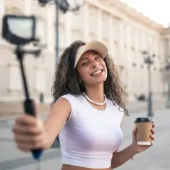 Uśmiechnięta content creator z kawą w ręku nagrywa filmik na selfie sticku przed piękną budowlą.