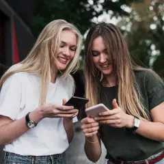Dwie przyjaciółki śmieją się, przeglądając telefony. Jedna z nich pokazuje drugiej, jak założyć drugie konto na Facebooku.