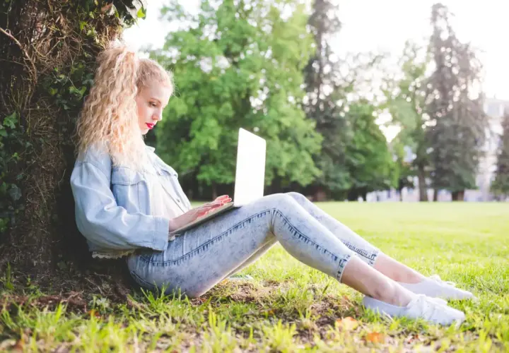 Dziewczyna z blond lokami siedzi pod drzewem i pisze na laptopie. Może właśnie szuka inspiracji, jak zacząć bloga.