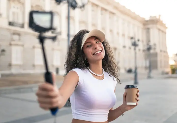 Uśmiechnięta content creator z kawą w ręku nagrywa filmik na selfie sticku przed piękną budowlą.