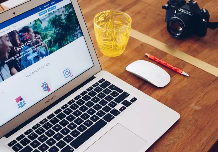 Laptop z ekranem pokazującym jak zrobić post sponsorowany na Facebooku, obok myszka, długopis i aparat.