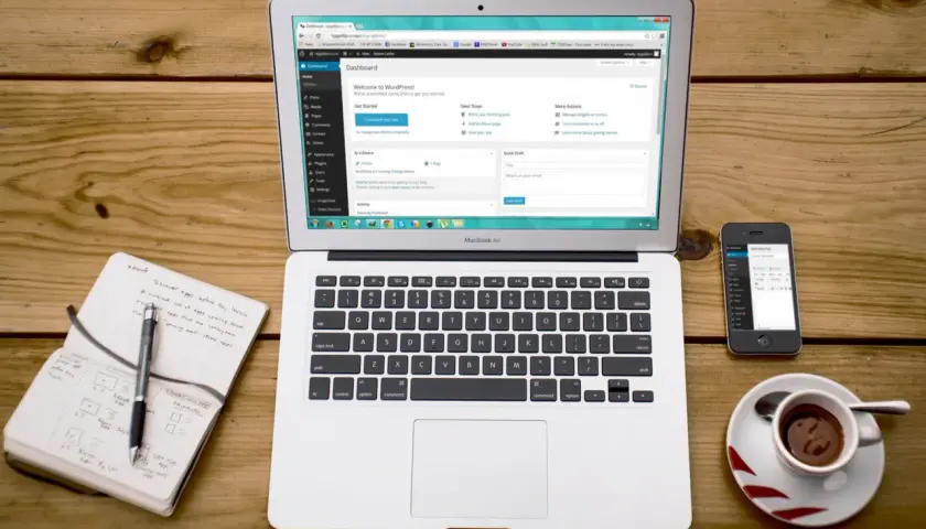 Laptop z panelem WordPress, notatnik, smartfon i filiżanka kawy na drewnianym stole.
