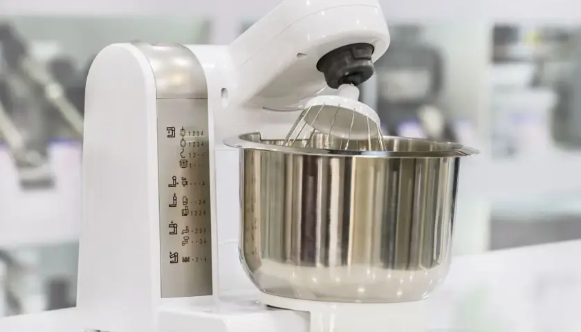 Biały robot kuchenny z metalową misą i trzepaczką. Na obudowie widoczne ikony funkcji, pomocne przy wyborze, jaki robot kuchenny wybrać blog.