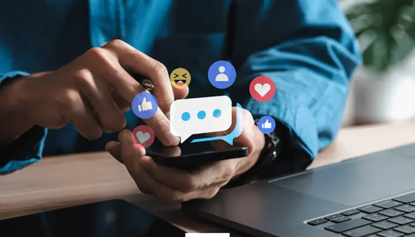 Ręce użytkownika na smartfonie, otoczone ikonami reakcji z Facebooka. Dowiedz się, jak wyróżnić post na Facebooku, by zdobyć więcej zaangażowania.