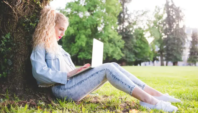 Dziewczyna z blond lokami siedzi pod drzewem i pisze na laptopie. Może właśnie szuka inspiracji, jak zacząć bloga.