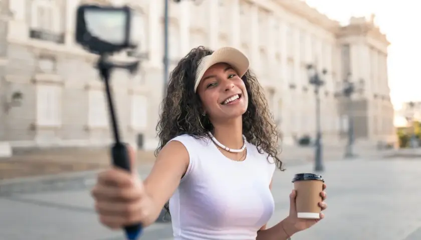 Uśmiechnięta content creator z kawą w ręku nagrywa filmik na selfie sticku przed piękną budowlą.
