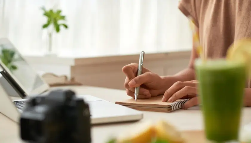 Osoba planuje, jak zarabiać na blogu kulinarnym, notując pomysły przy laptopie, z aparatem i zielonym koktajlem.