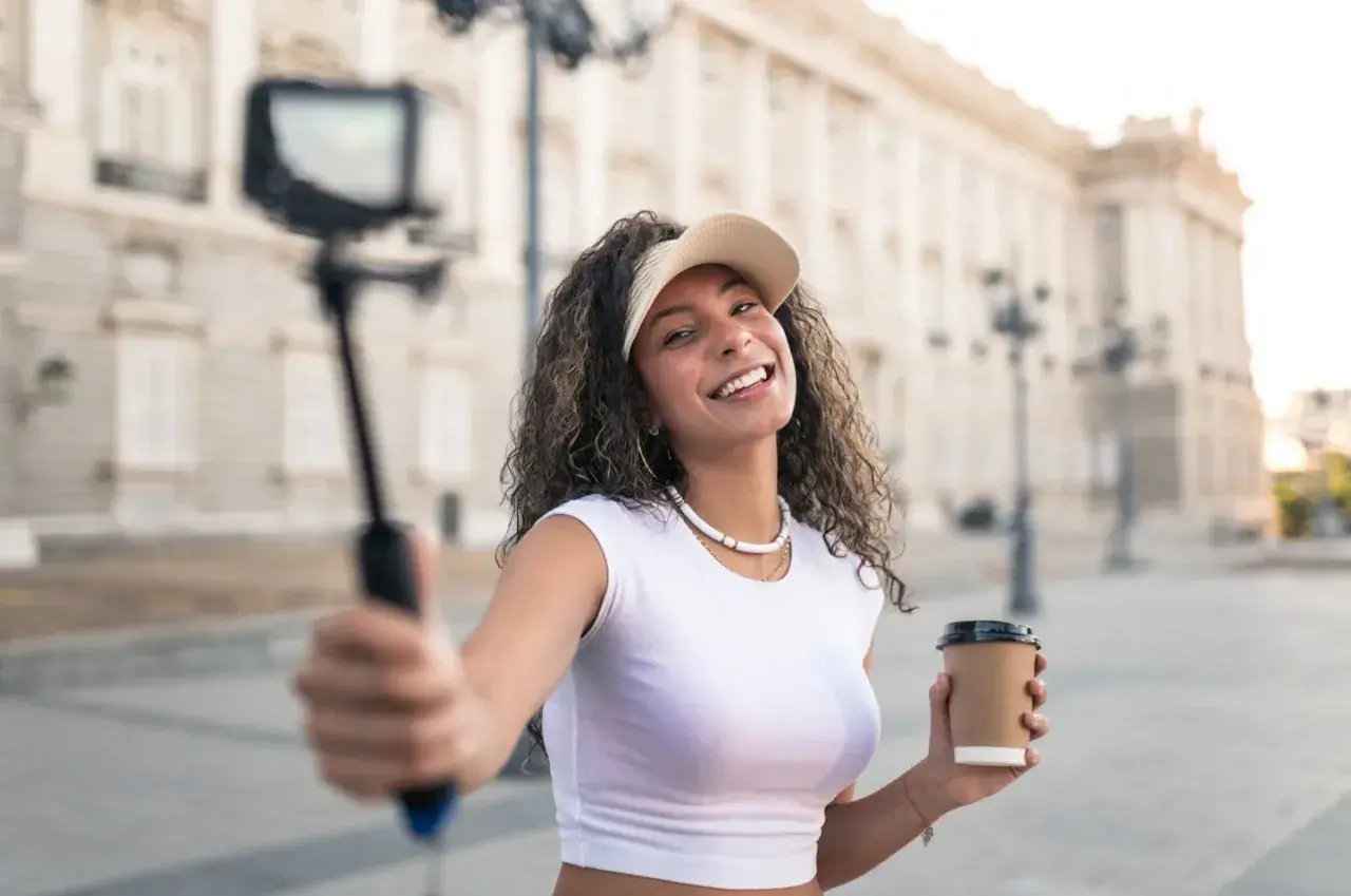 Uśmiechnięta content creator z kawą w ręku nagrywa filmik na selfie sticku przed piękną budowlą.