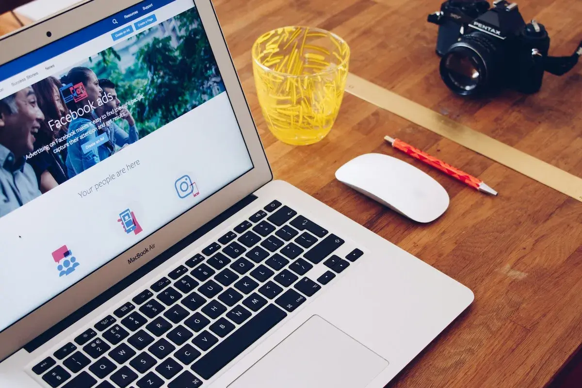 Laptop z ekranem pokazującym jak zrobić post sponsorowany na Facebooku, obok myszka, długopis i aparat.