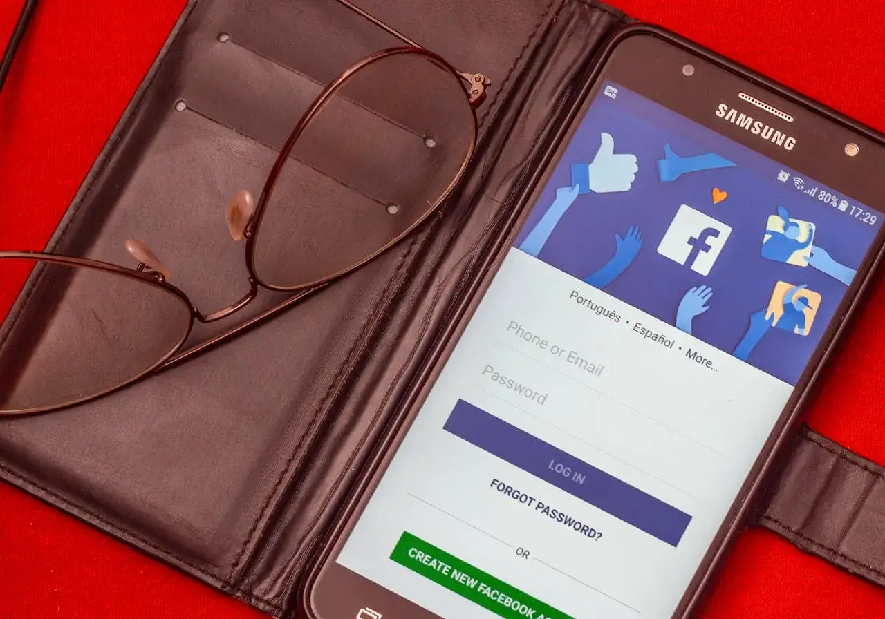 Okulary leżą na etui, obok smartfon z ekranem logowania Facebooka. Zobacz, jak dodawać zdjęcia na Facebooku.
