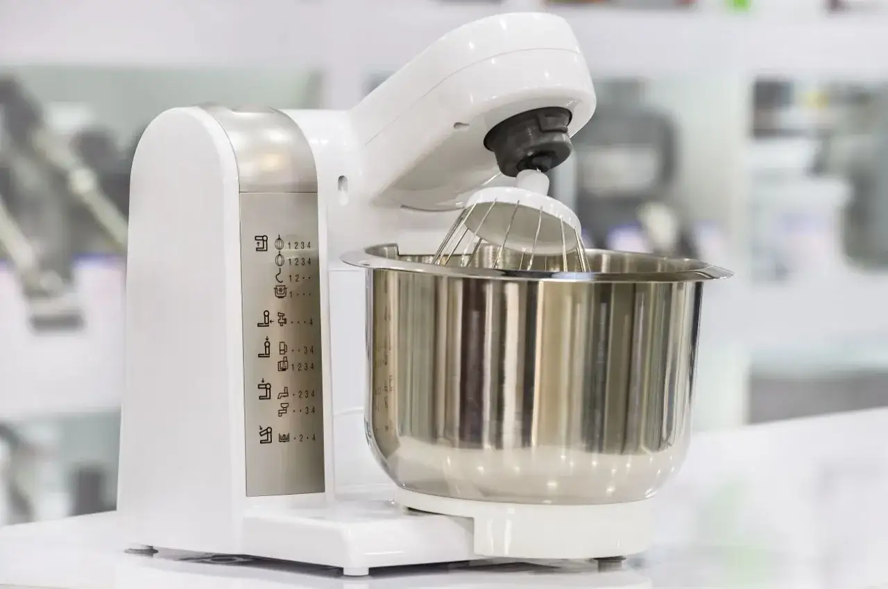Biały robot kuchenny z metalową misą i trzepaczką. Na obudowie widoczne ikony funkcji, pomocne przy wyborze, jaki robot kuchenny wybrać blog.
